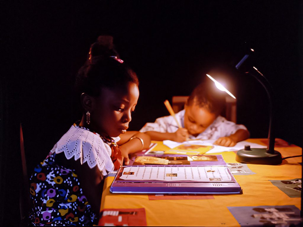 Two children studying and doing homework at night under a table lamp in a dark setting, education photography in Nigeria
