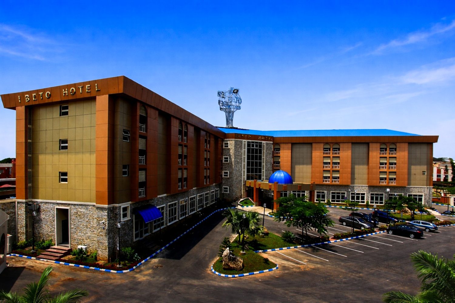 Wide panoramic view of Ibeto Hotel showcasing its modern architecture, entrance, and parking area during the day.