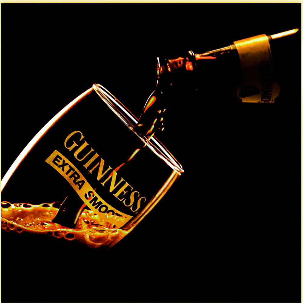 Guinness Extra Smooth beer bottle pouring into a tilted branded glass with golden foam against a black background.